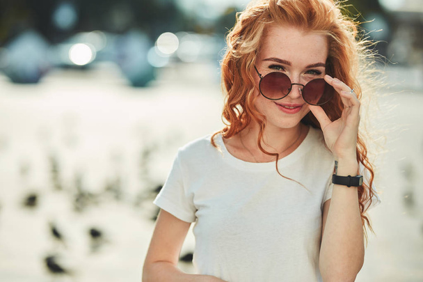Beautiful red-haired girl having fun on the street. The girls have a beautiful figure, a white T-shirt and jeans with sunglasses - Photo, Image