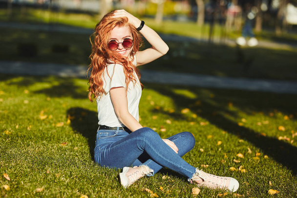Güzel bir kızıl saçlı kız, güneş ışınları tarafından ısınmış, bir çim üzerinde oturuyor ve kamera için poz. Kız kot pantolon, yüzünde gözlük ve kolunda modern bir alet olan bir tişört giyiyor. - Fotoğraf, Görsel