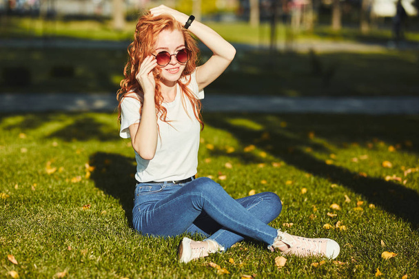 太陽の光に温められた美しい赤毛の少女が芝生の上に座り、カメラのポーズをとっている。少女はジーンズ、メガネ、腕にモダンなガジェットをつけたTシャツを着ている。 - 写真・画像