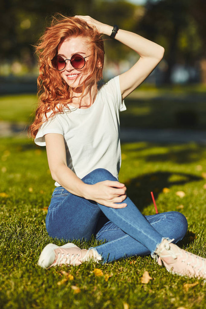 Una encantadora chica pelirroja, calentada por los rayos del sol, está sentada en un césped y posando para la cámara. La chica lleva una camiseta con jeans, gafas en la cara y un gadget moderno en el brazo.
 - Foto, imagen