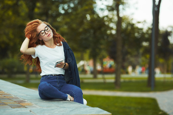 Güzel bir kızıl saçlı kız parkta oturuyor ve kamera için poz. Kız kot pantolon, yüzünde gözlük ve kolunda modern bir alet olan bir tişört giyiyor. - Fotoğraf, Görsel