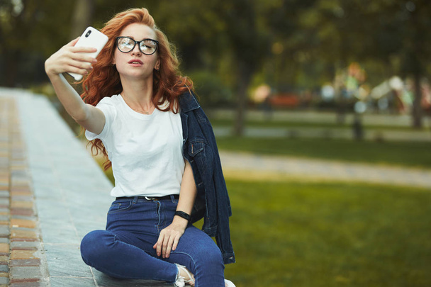Una bella ragazza dai capelli rossi si riposa in un giardino pubblico e si fa un selfie sul suo cellulare. La ragazza indossa una t-shirt con jeans, occhiali sul viso e un gadget moderno sul braccio.
 - Foto, immagini