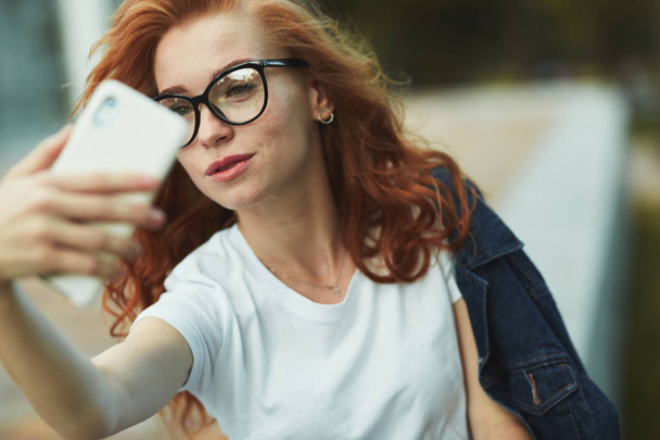 A beautiful red-haired girl is resting in a public garden and takes a selfie on her mobile phone. The girl is wearing a T-shirt with jeans, glasses on her face, and a modern gadget on her arm - Foto, afbeelding