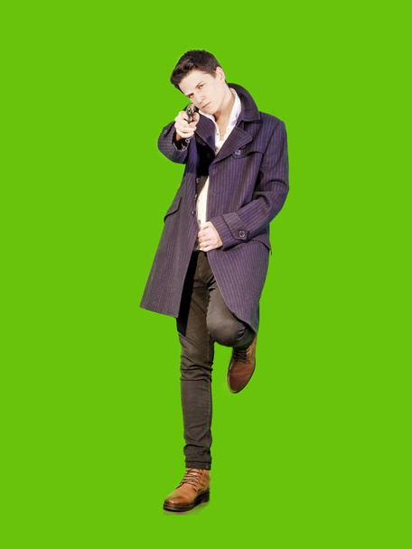 Full length of a focused thoughtful man in a black coat, standing and holding a gun on a green background - Foto, Imagen