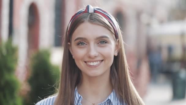 Portrait of a beautiful young woman looking into the camera and smiling standing on the old street background. Girl wearing in striped shirt dress with headband. - Filmati, video