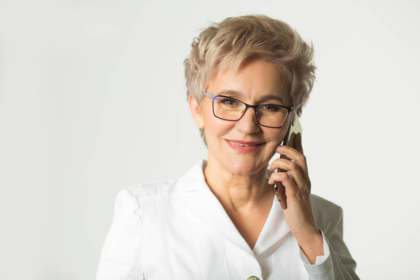 portrait of an adult woman with a short haircut wearing glasses in a white jacket on a white background makes phone calls - Fotó, kép