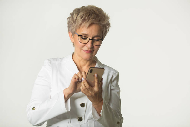 portrait of an adult woman with a short haircut wearing glasses in a white jacket on a white background with a phone in her hand - Photo, Image