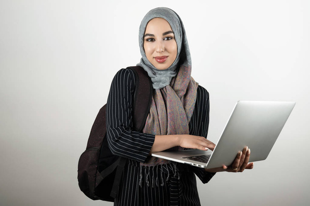 young attractive Muslim student with bagpack holding and tapping her laptop wearing turban hijab headscarf isolated white background - Foto, afbeelding