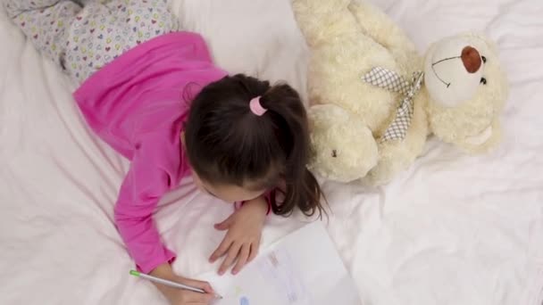 χαριτωμένο μικρό κορίτσι σχεδιάζοντας φωτογραφίες, ενώ ξαπλωμένο στο κρεβάτι. - Πλάνα, βίντεο