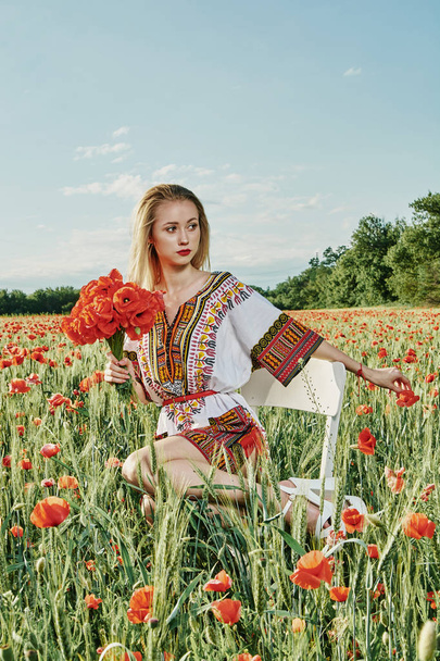 Long-haired blonde young woman in a white short dress on a field of green wheat and wild poppies. - Photo, Image