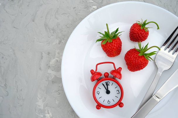 Red ripe strawberries berry on white plate, cutlery and red alarm clock on gray stone background table, closeup. Top view, flat lay, copy space. Concept diet and detox time or summer menu time - Фото, зображення
