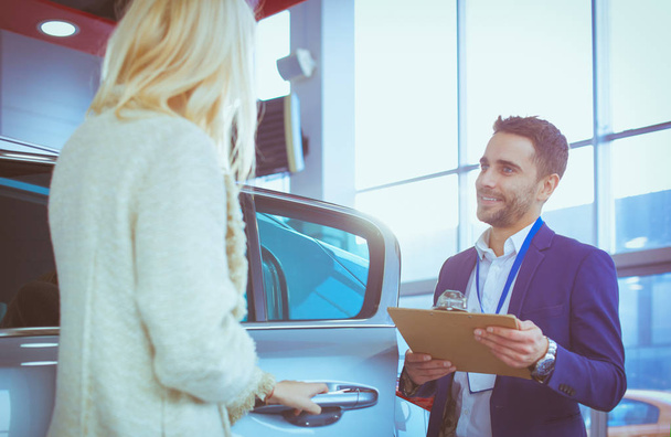 Beautiful young woman is talking to handsome car dealership worker while choosing a car in dealership - 写真・画像