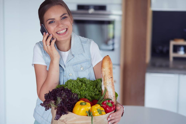 Smiling woman with mobile phone holding shopping bag in kitchen - Valokuva, kuva