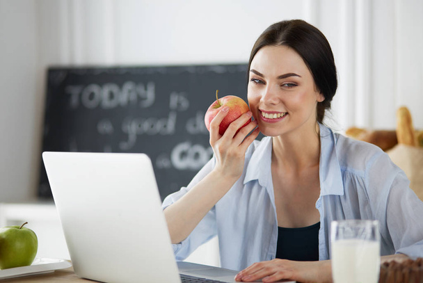 Woman with apple and laptop sitting in the kitchen - Photo, image