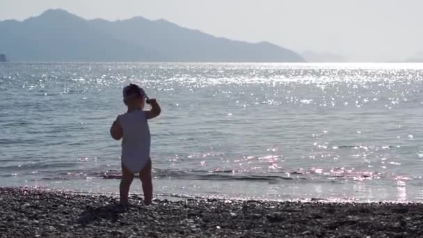 Rozkošný batole, který stojí zády ke kameře, hází oblázek do moře - Záběry, video