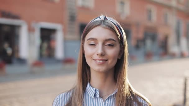 Portrait of a beautiful young woman looking into the camera and smiling standing on the old street background. Girl wearing in striped shirt dress with headband. - Felvétel, videó