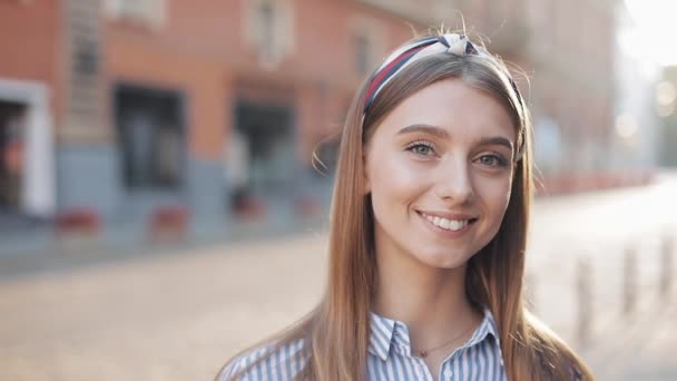 Portrait of a beautiful young woman looking into the camera and smiling standing on the old street background. Girl wearing in striped shirt dress with headband. - Кадры, видео