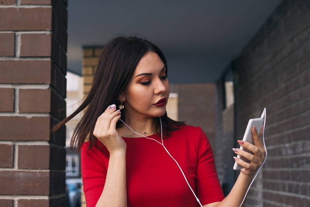 beautiful young woman with red lipstick in red dress uses phone and earphones - Fotografie, Obrázek