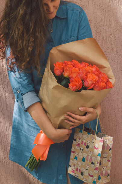 young woman in denim dress with bouquet of roses and gift paper bag - Fotó, kép