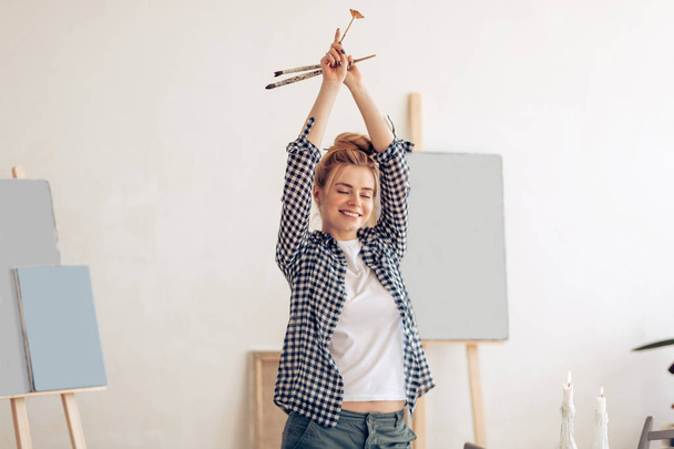 cheerful funny girl with hairbun raising her arms and holding brushes - Foto, imagen