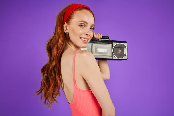 red-haired girl with freckles stands with portable audio player, looks playfully in camera - Photo, image