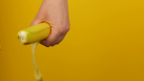 Female hand with yellow manicure peels the skin a ripe banana fruit on yellow background - Кадри, відео