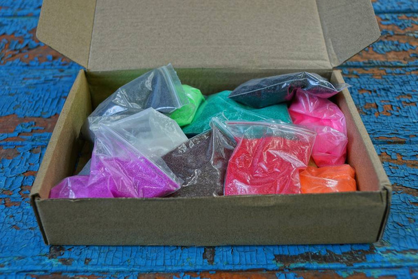pile de sacs en plastique avec du sable coloré dans une boîte en papier ouverte se tient sur une table bleue
 - Photo, image