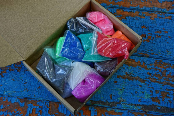een stapel plastic zakken met gekleurd zand in een open papier doos staat op een blauwe tafel - Foto, afbeelding