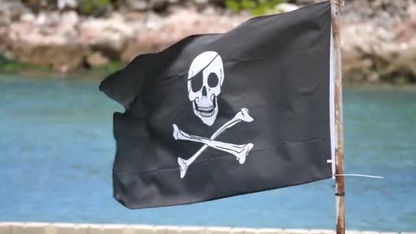 A black and white pirate skull and bones flag mounted to a weathered and rusted steel post flaps in the wind with blue water and rocky shoreline in the background. - Footage, Video