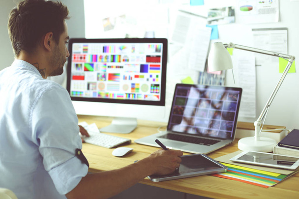 Retrato del joven diseñador sentado en el estudio gráfico frente a la computadora portátil y el ordenador mientras trabaja en línea. - Foto, imagen