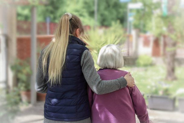 junge Pflegerin geht mit der älteren Frau im Garten mit hellem Hintergrund - Foto, Bild