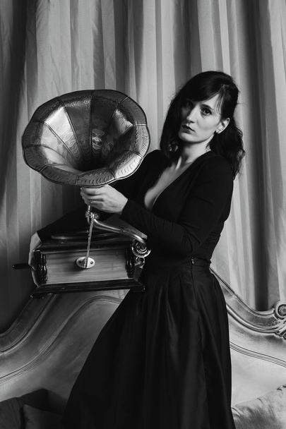 Black and white portrait of young beautiful female model in black sexy dress standing on bed in with the gramophone interior studio. - Fotó, kép