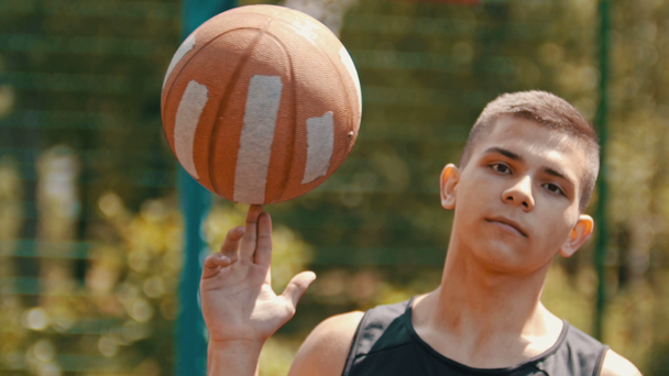 Ένα έφηβο αγόρι στέκεται σε ένα αθλητικό έδαφος και γυρίζει την μπάλα του μπάσκετ στο δάχτυλό του - Πλάνα, βίντεο