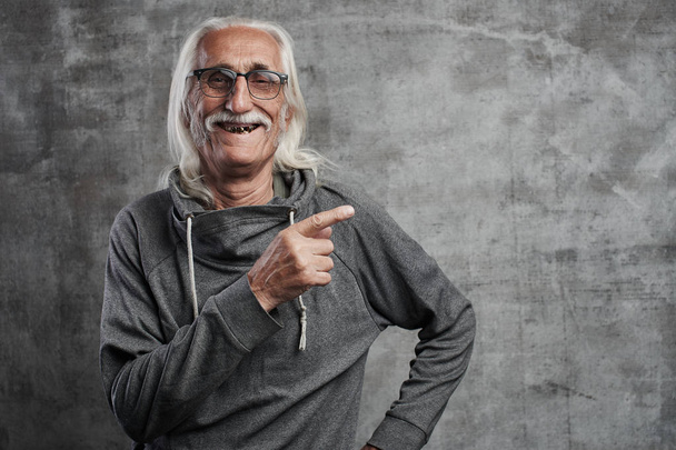 Aged gray haired smiling Caucasian man points to the side with index finger, advertises something. Grandfather in glasses with mustache recommends buying your product  - Photo, image