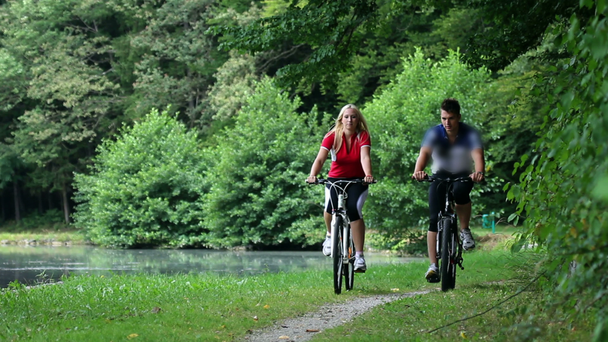 若いカップルがカメラに向かって湖の近くの自然の中でサイクリング - 映像、動画