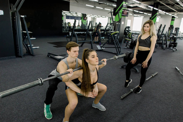 Attractive woman and handsome muscular man trainer are training with barbells in light modern gym. Beautiful girl watching at the workout - Fotó, kép