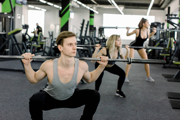Young people working out with barbells at gym. Attractive women and handsome muscular man trainer are training in light modern gym. Beautiful girl squats under the supervision of the coach. - Foto, Bild