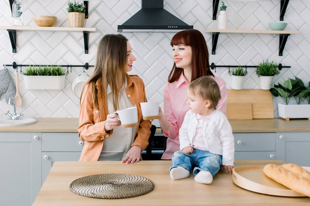 Two Cheerful young Women sitting at a table with cups in a kitchen. Little baby girl is sitting on the table. Breakfast, friendship concept - Photo, Image