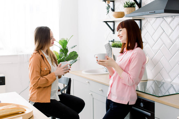 Portrait of two smiling women holding cups of coffee at home in the kitchen - Foto, Bild