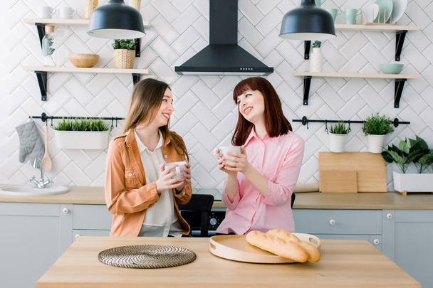 Two brunette girls friend drinking coffee or green tea talking. Couple of woman eating breakfast together on kitchen interior background - 写真・画像
