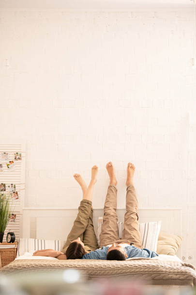 Young carefree barefoot dates in casualwear lying on bed with their raised legs opposite wall while relaxing at home - Fotoğraf, Görsel