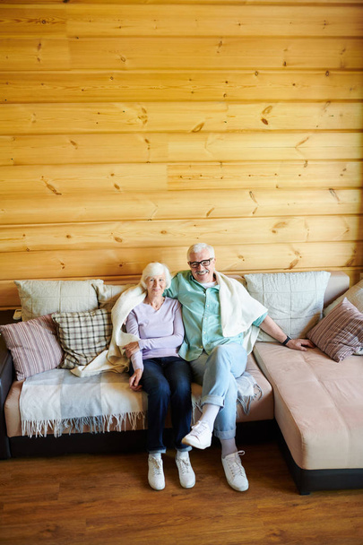 Affectionate senior couple in casualwear relaxing on sofa in their country house on background of wooden wall - Фото, изображение