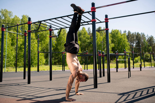 Attractive young muscular athlete doing handstand on sportsground on sunny day with facilities on background - Photo, image