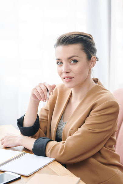 Portrait of content confident brunette business lady in beige jacket sitting at table and keeping list of tasks - Фото, изображение