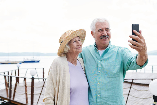 陽気な先輩夫婦が夏の日に水辺を背景にスマートフォンで自分撮りをする - 写真・画像