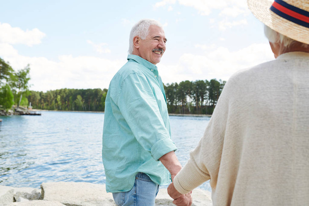 Heureux homme âgé en tenue décontractée tenant sa femme à la main tout en passant le week-end par le lac à la campagne
 - Photo, image