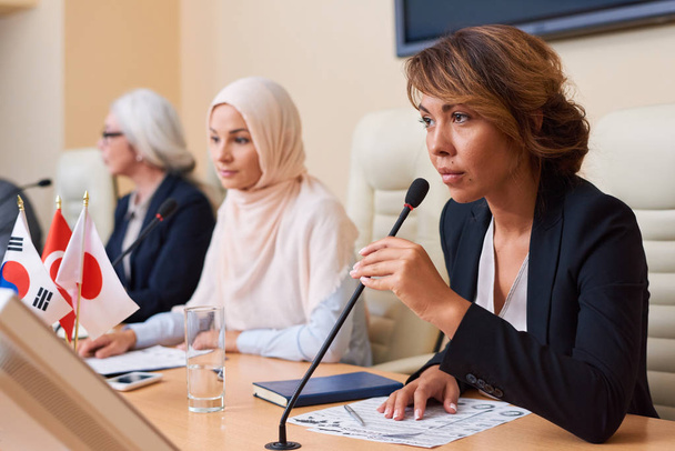 Молодая уверенная в себе женщина в формальной одежде держит микрофон у рта, когда собирается ответить или задать вопрос на конференции
 - Фото, изображение