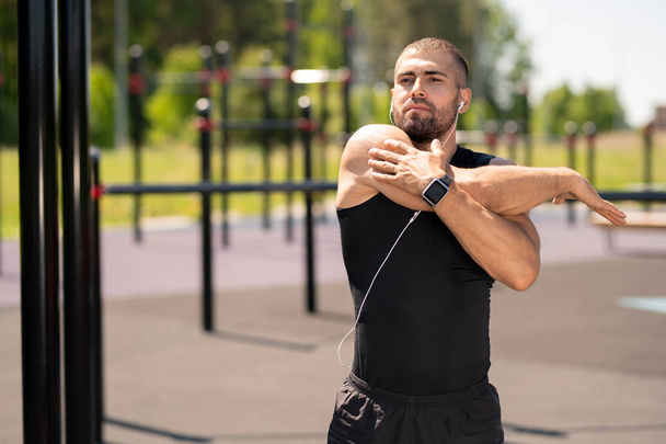 Young muscular athlete in tracksuit stretching right arm while exercising on background of sports facilities - Фото, изображение