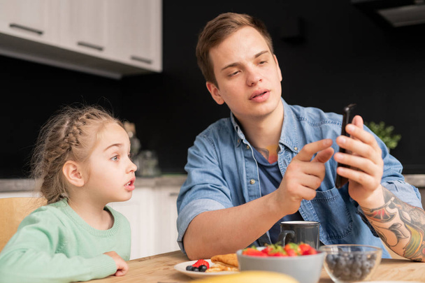 Serious handsome hipster father using smartphone and showing internet video to daughter while they having breakfast in kitchen - Fotó, kép