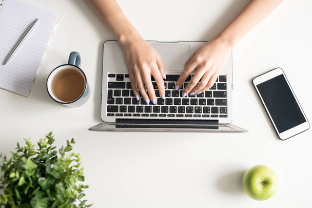 Vista superior de la joven freelancer manos sobre el teclado portátil pf durante la red con manzana, taza de té y teléfono inteligente cerca de
 - Foto, Imagen